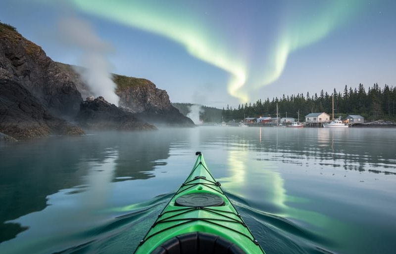 Kayak on calm coastal water near dark cliff geothermal pools, with misty treeline and faint northern lights in the evening sky.